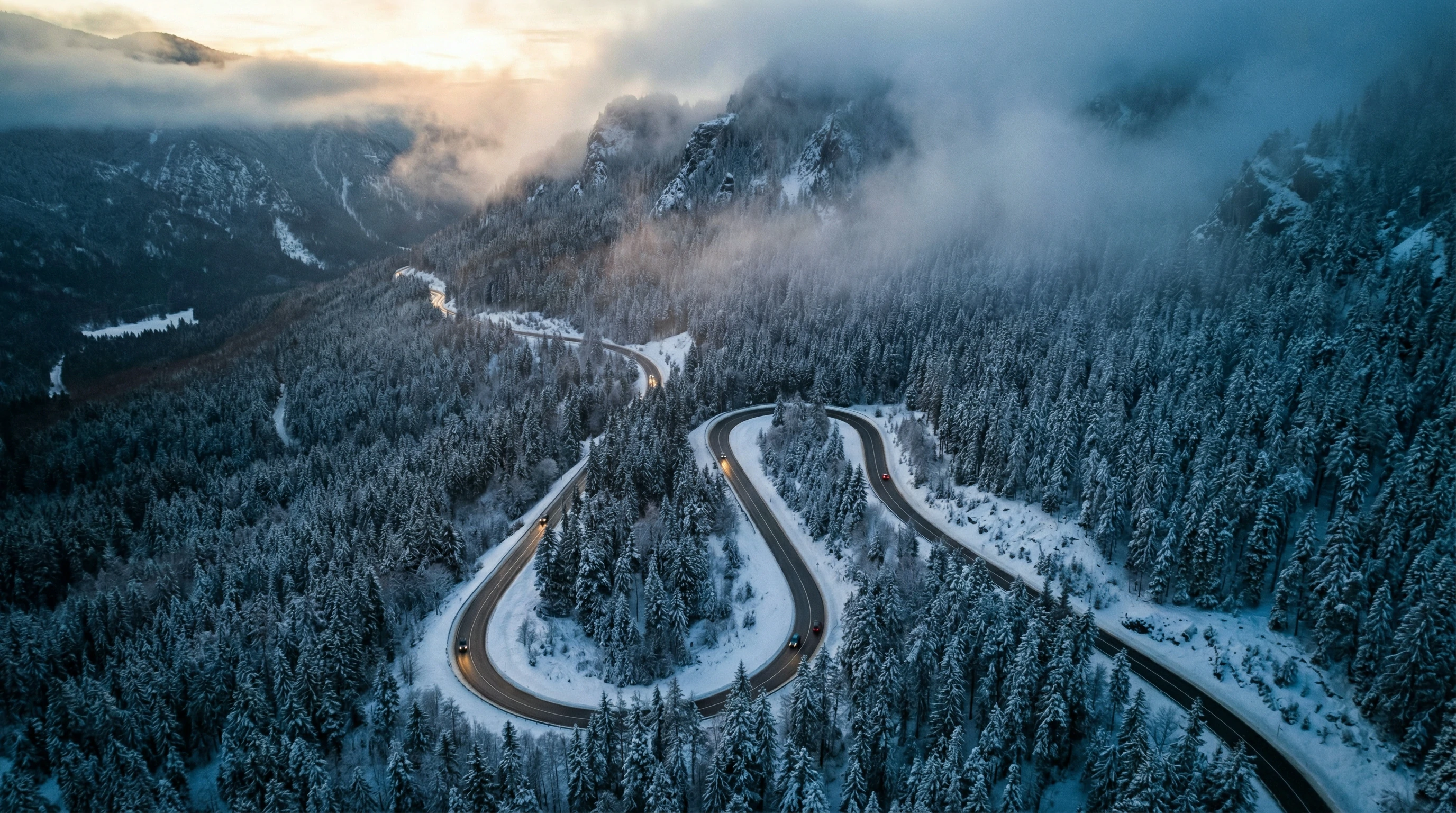 Aerial drone shot of a winding hairpin road cutting through a dense, snow-covered evergreen forest. Cars navigate the curvy mountain pass under heavy fog and a soft winter sunrise.